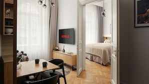A modern hotel room with a wooden herringbone floor, a dining area with a table and chairs, a TV mounted on the wall, and a bed with a beige blanket. - R16 Residences Prague (Prague)
