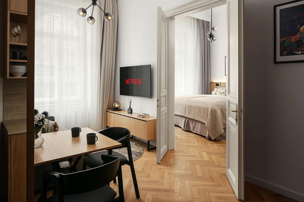 Una habitación de hotel moderna con suelo de madera en espiga, una zona de comedor con mesa y sillas, un televisor montado en la pared y una cama con una manta color beige.