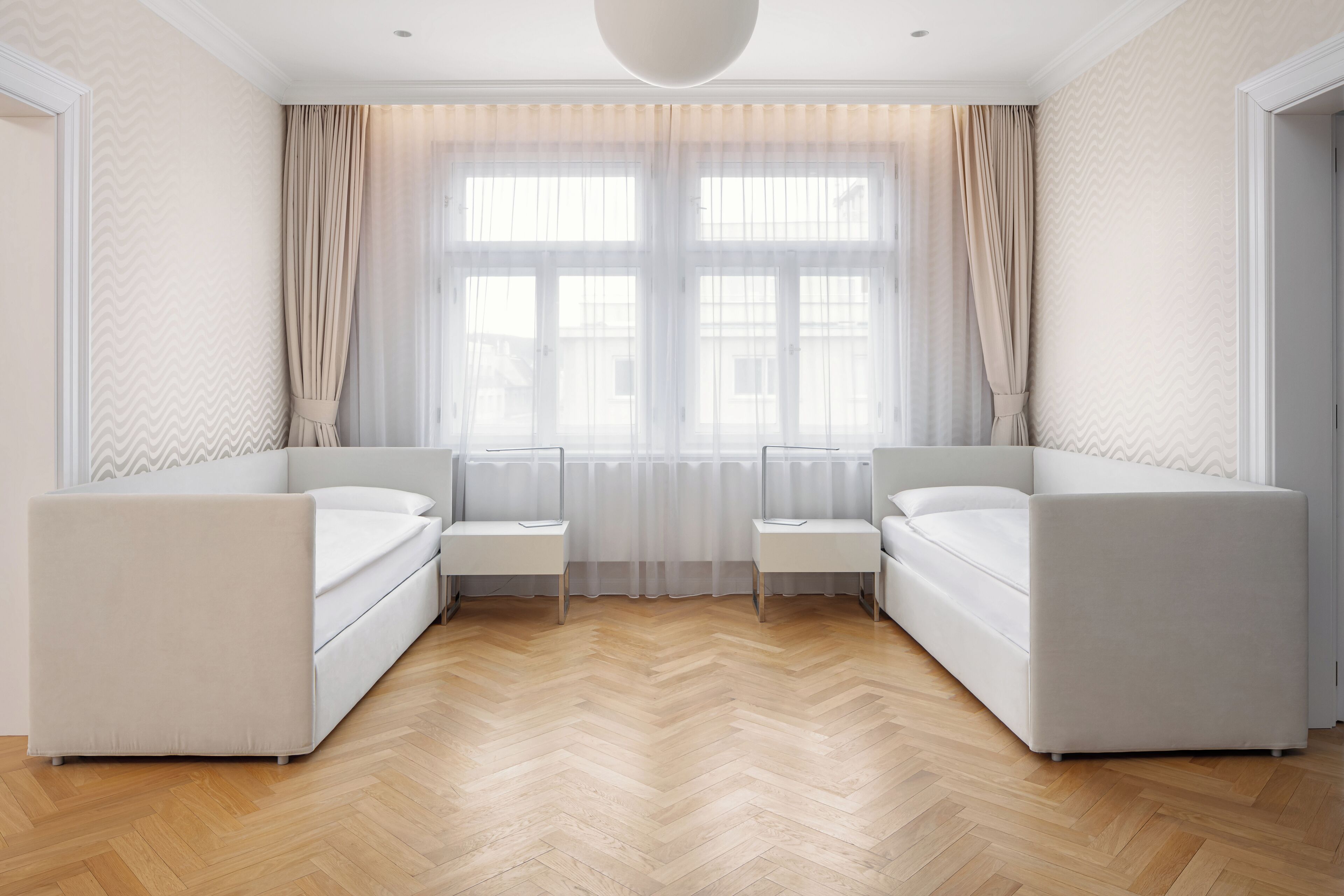 Quarto de hotel moderno com uma janela grande, dois sofás brancos, uma mesa pequena e um chão de madeira em padrão herringbone.