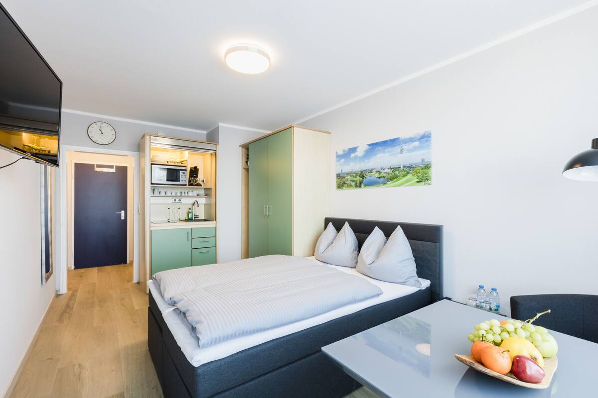 Une chambre à coucher moderne avec un lit, une table de chevet sur laquelle se trouvent des fruits, une vue sur une cuisine et un tableau accroché au mur.