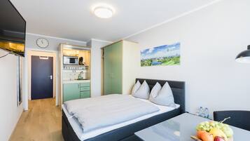 A modern bedroom with a bed, a bedside table with fruit, a view of a kitchen, and a painting on the wall.