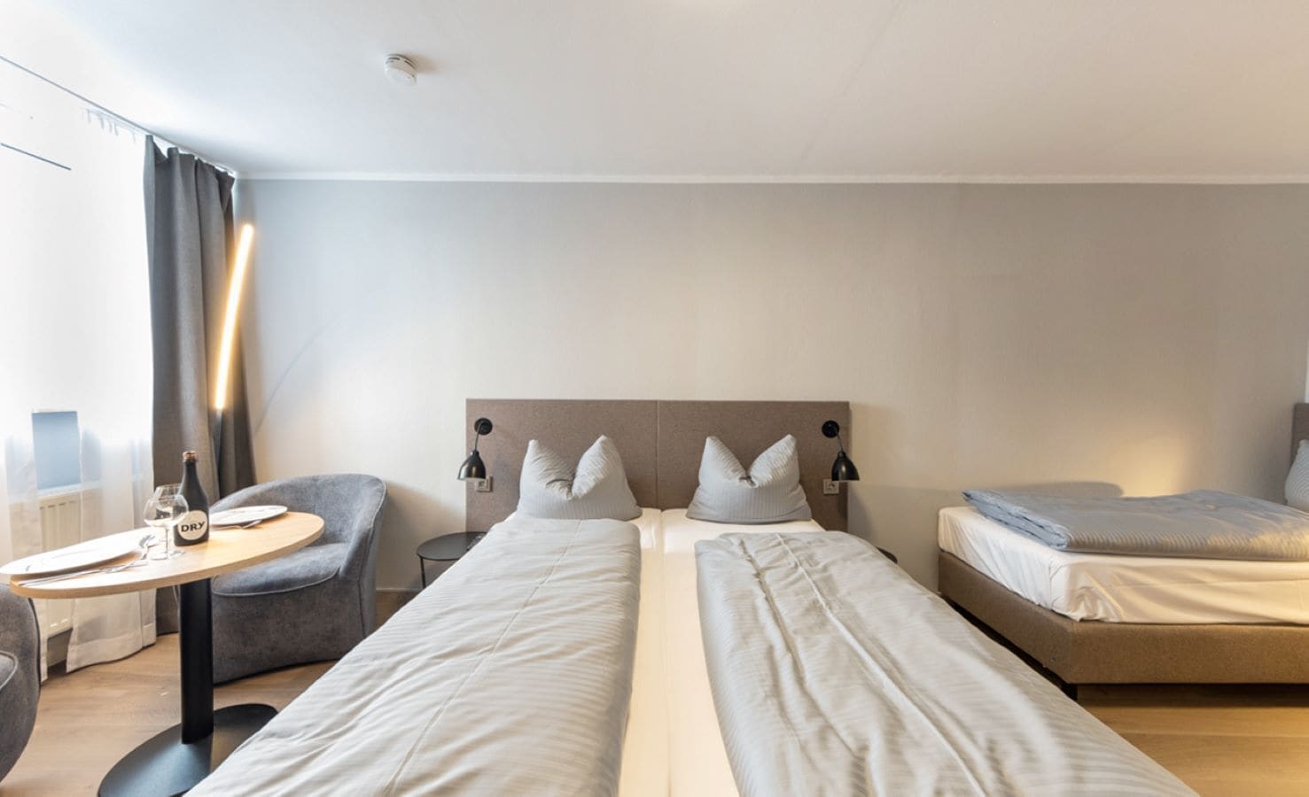 A hotel room with two beds, a round table with a bottle and glasses, a chair, and a nightstand with a lamp.