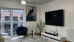 Een moderne woonkamer met een groot raam, een blauwe fauteuil, een glazen salontafel, een witte bank, een wandtelevisie en een vloerlamp.