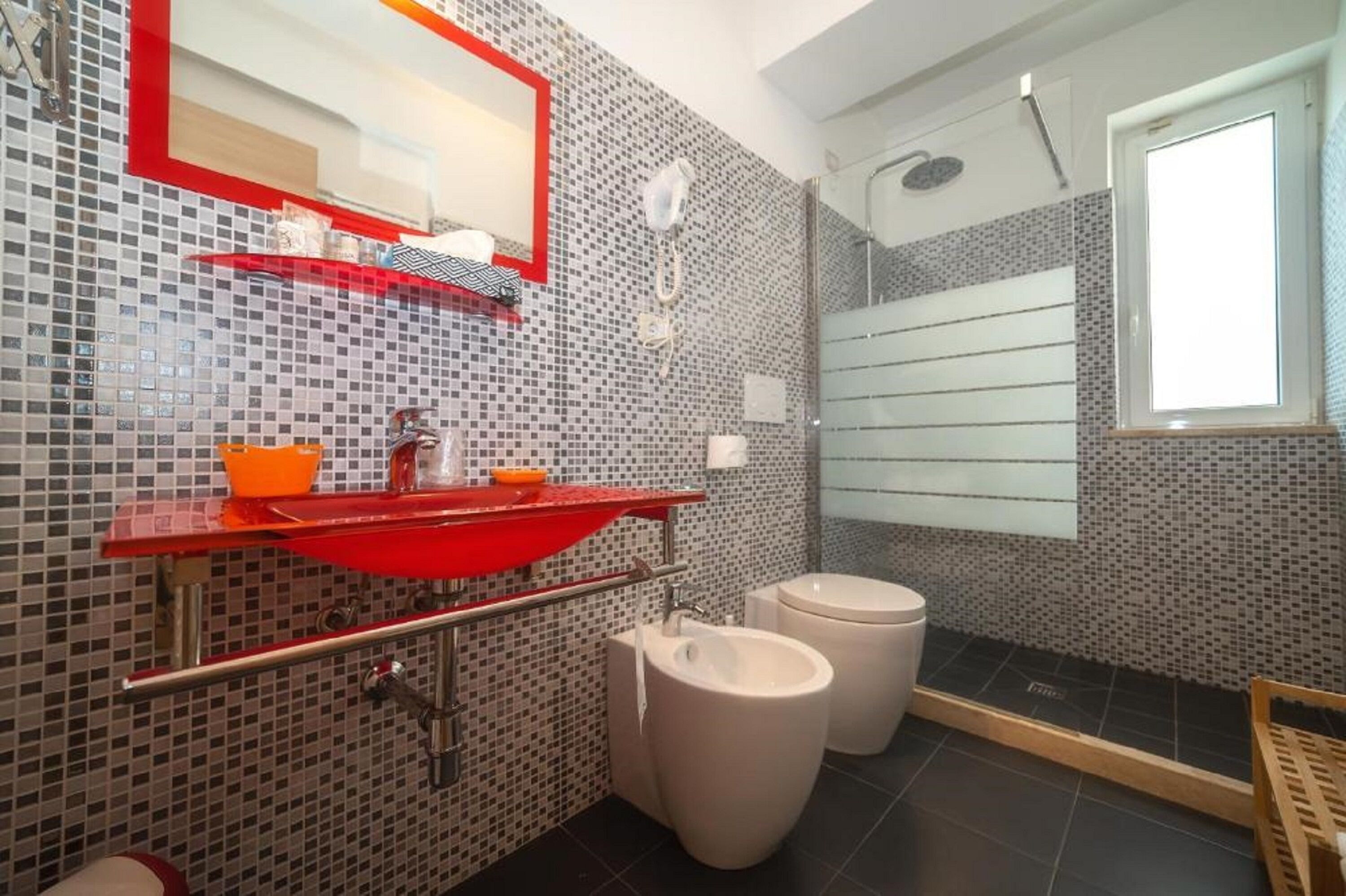 A bathroom with a red sink, two toilets, a shower, and a mirror with a red frame.