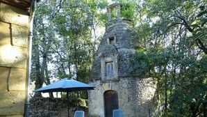 Ein rustikales Steinhaus mit Schornstein, umgeben von Bäumen. Ein Picknicktisch mit blauem Sonnenschirm und zwei Stühlen steht daneben.