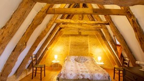 Ein gemütliches Schlafzimmer im Dachgeschoss mit schrägen Wänden, Holzbalken, einem Bett mit floralem Bettbezug, zwei Nachttischen und einem kleinen Hocker.