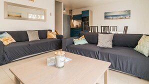 Un salon moderne avec deux canapés gris, une table basse en bois et une petite bougie. On aperçoit une cuisine à l’arrière-plan, avec des meubles bleus, ainsi qu’un coin repas avec des chaises.