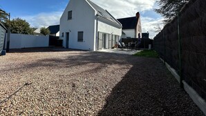 Un bâtiment blanc avec un toit en pente, une allée gravillonnée et un espace clos aménagé avec du mobilier d’extérieur.