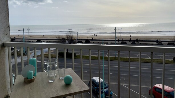 A balcony with a table, a view of the beach, and a parking lot.