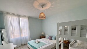 A bedroom with a bed, a desk, a chair, a bedside table, and a hanging light fixture.