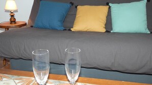 A bed with grey bedding, three pillows in blue, yellow, and teal, and two clear glasses on a table.
