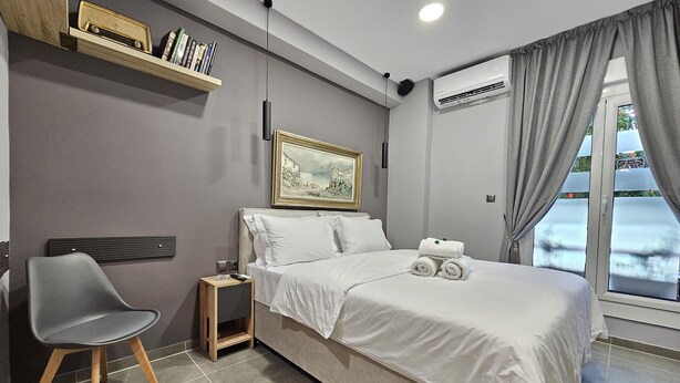 A modern bedroom with a bed, a chair, a nightstand, a shelf with books, a painting, and an air conditioning unit. - "Dione" Luxury Business Suite in the Heart of Athens (Athina)