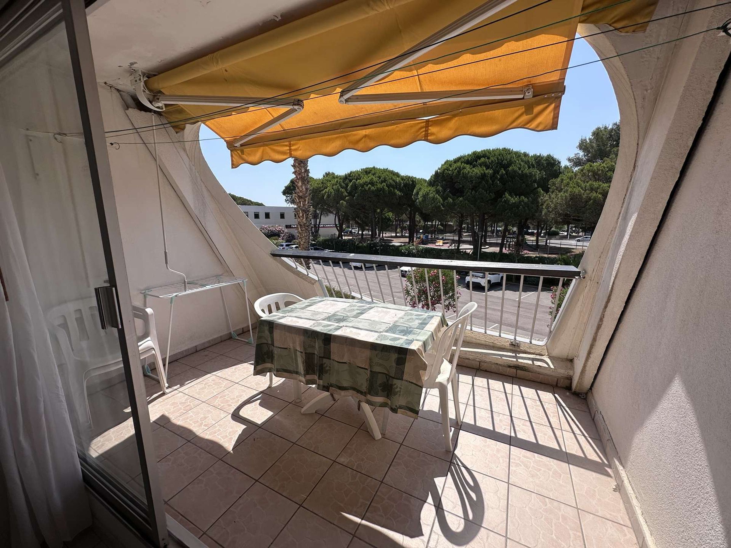 Un balcone con tavolo e sedie, un tendone giallo e vista su alberi e un parcheggio.
