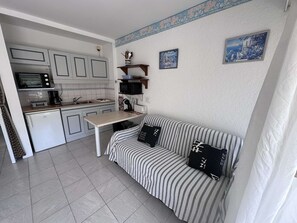Eine kompakte Küche mit Mikrowelle, Toaster und kleinem Kühlschrank. Ein gestreiftes Sofa mit schwarzen Kissen lehnt an der Wand, darüber hängen zwei kleine gerahmte Bilder.