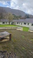 Un patio con zona pavimentada, una carretilla y una hilera de edificios con una montaña al fondo.