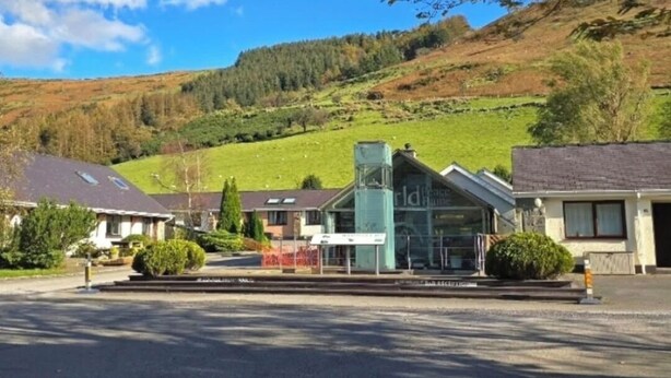 Un hotel con fachada de vidrio moderna, rodeado por un estacionamiento y una vista panorámica a una colina.