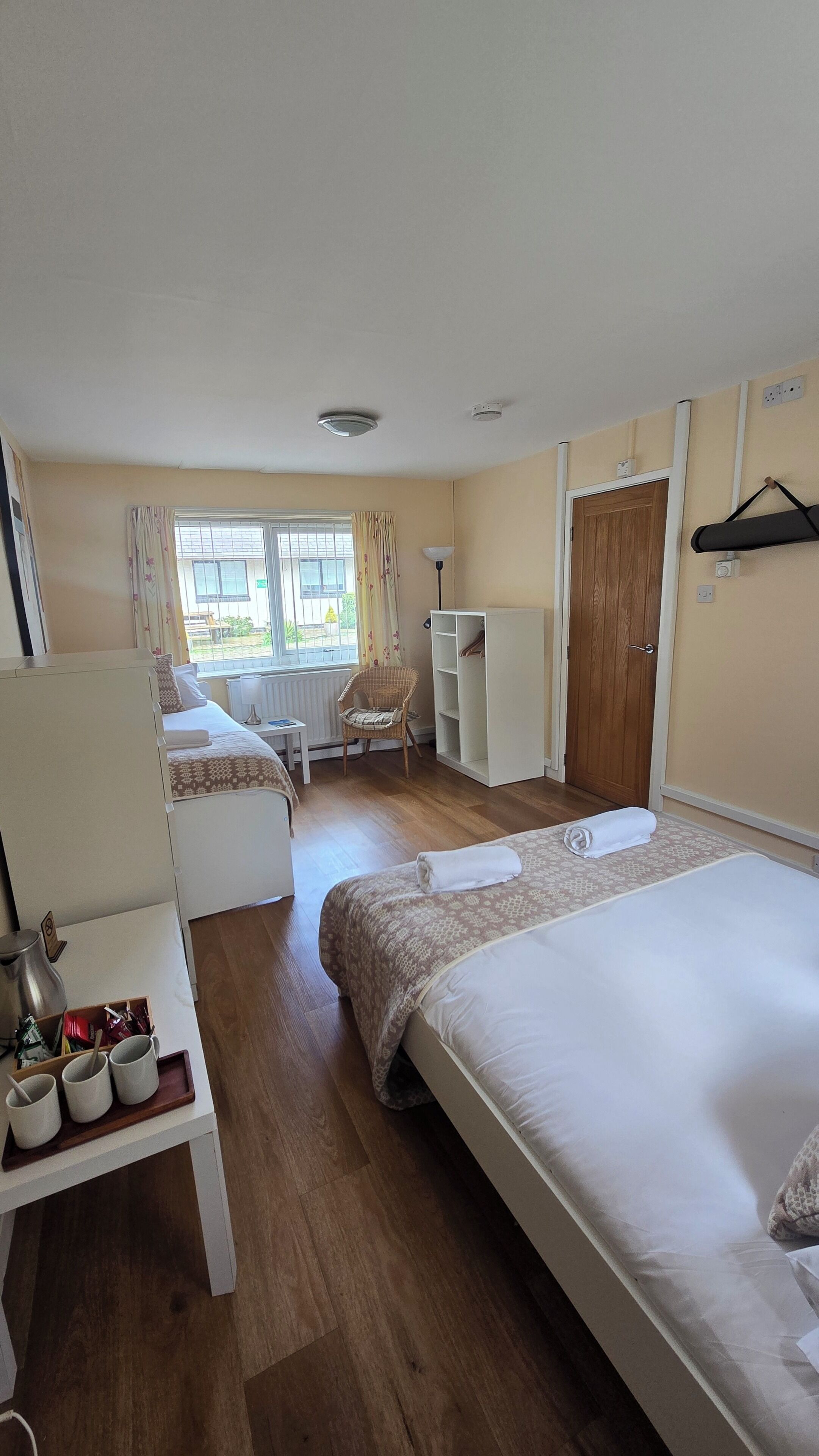 A hotel room with two beds, a desk with a tray of mugs, a chair, a wardrobe, and a window with curtains.