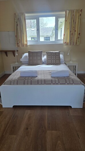 A neatly made bed with patterned bedding, a bedside table with a lamp, and a window with curtains. - Snowdonia Mountain Lodge (Bangor)