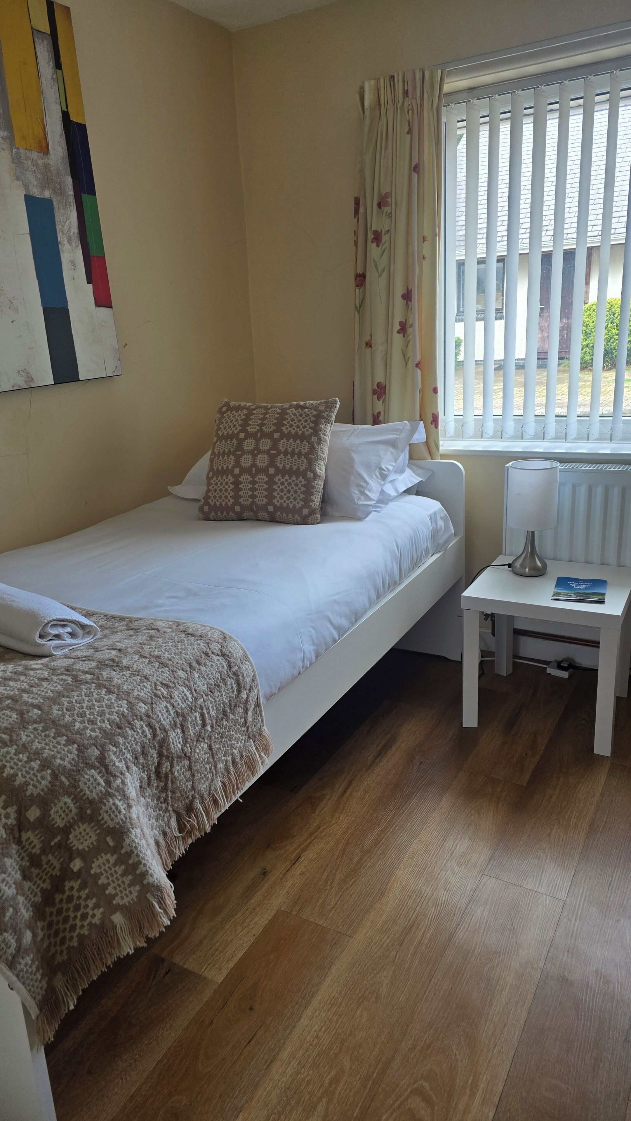 A single bed with a patterned pillow, a bedside table with a lamp, and a window with blinds.