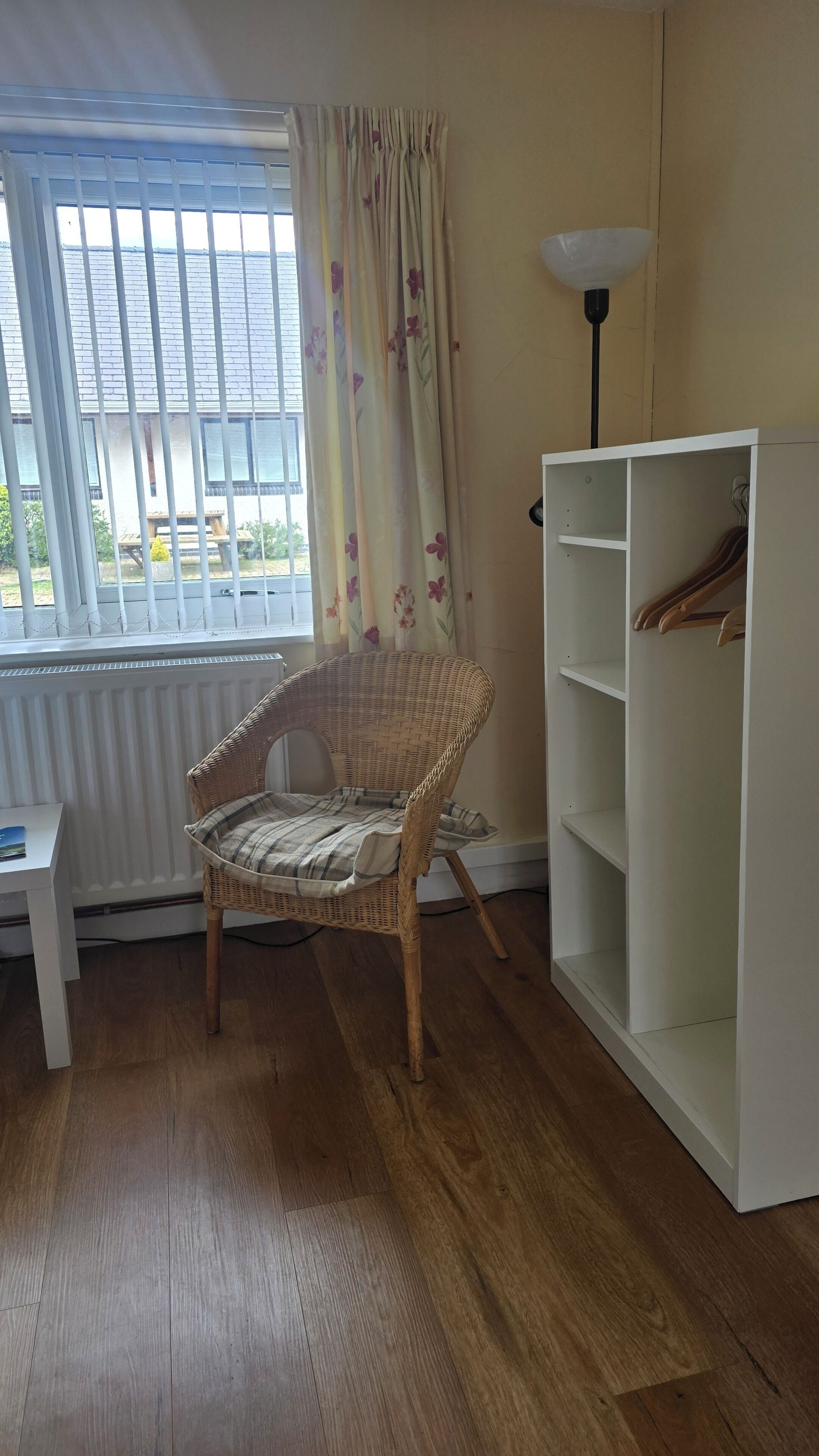 A room with a wicker chair, a white shelving unit, a radiator, and a window with blinds.