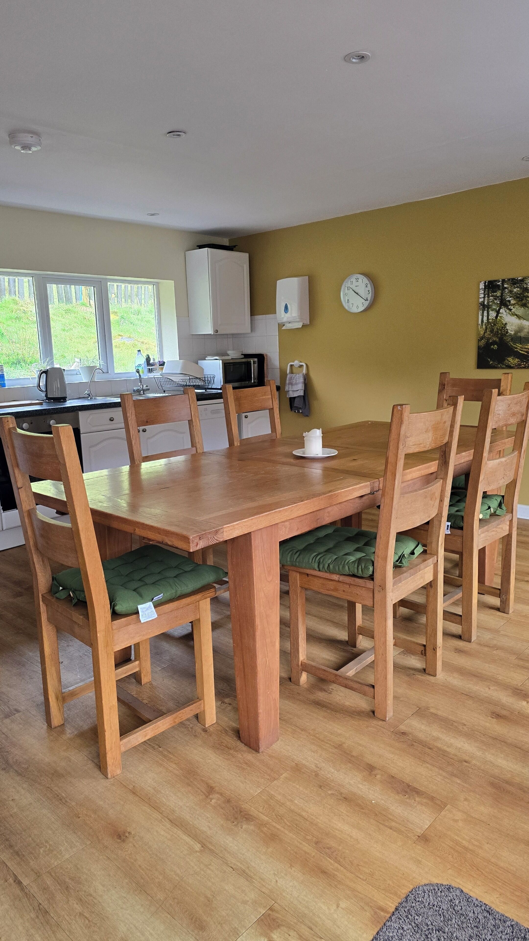 Un comedor con una mesa y sillas de madera, un reloj en la pared y una cocina al fondo.