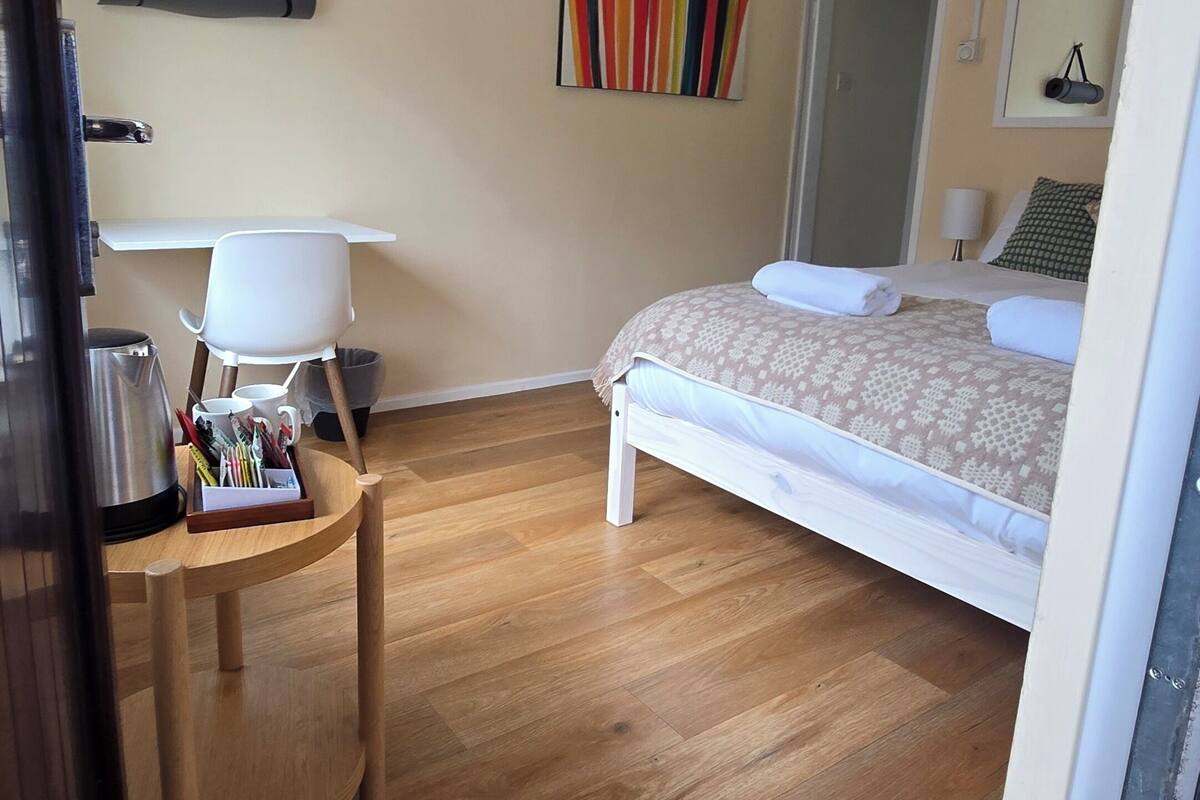 Habitación de hotel con cama, escritorio y silla, mesita con tetera y tazas, un cuadro llamativo en la pared y una puerta que da a otra habitación.