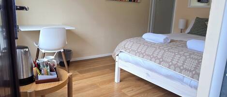 Habitación de hotel con cama, escritorio y silla, mesita con tetera y tazas, un cuadro llamativo en la pared y una puerta que da a otra habitación.