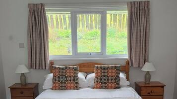 Una cama doble con ropa de cama estampada, dos mesitas de noche con lámparas, una ventana con cortinas y vistas al exterior con vegetación.