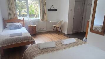 A hotel room with two beds, a desk, a chair, a radiator, a window with curtains, and a door.