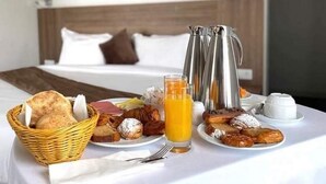 A breakfast setup with bread, pastries, and orange juice on a table in a hotel room with a bed and artwork on the wall.