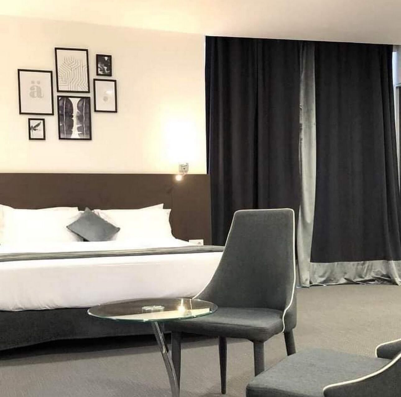 A hotel room with a bed, a chair, a small table, and a wall with framed pictures.