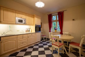 A kitchen with wooden cabinets, a microwave, a sink, and a checkered floor. There is a dining table with chairs and a window with red curtains. - Altes Pastorat: Wohnung mit Kamin und Großem Garten (Borrentin)