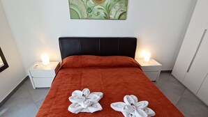 A neatly made bed with a red quilt and two white towels arranged in a flower shape on the bed. There are two bedside tables with lamps, and a piece of abstract art hangs on the wall above the bed. - Genius Home - Bright apartment in the heart of the historic center (Palermo)