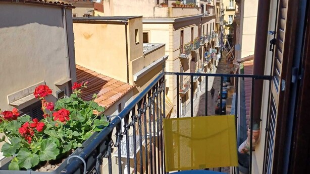 A narrow urban street with buildings on both sides, a balcony with red flowers, and a yellow chair. - Genius Home - Bright apartment in the heart of the historic center (Palermo)