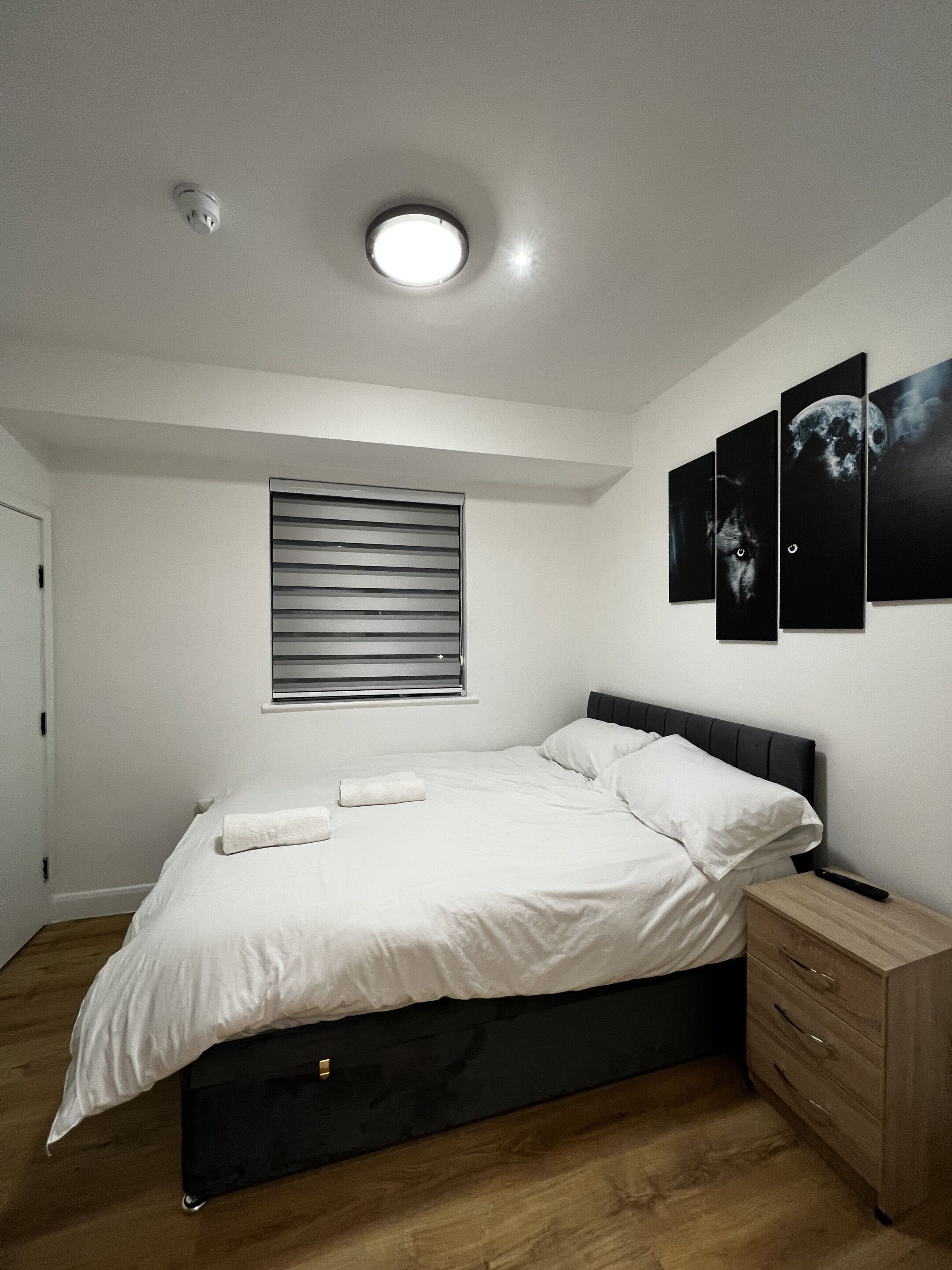 A bedroom with a bed, a bedside table, a window with blinds, and three wall-mounted pictures.