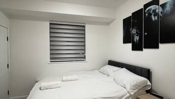 A bedroom with a bed, a bedside table, a window with blinds, and three wall-mounted pictures.