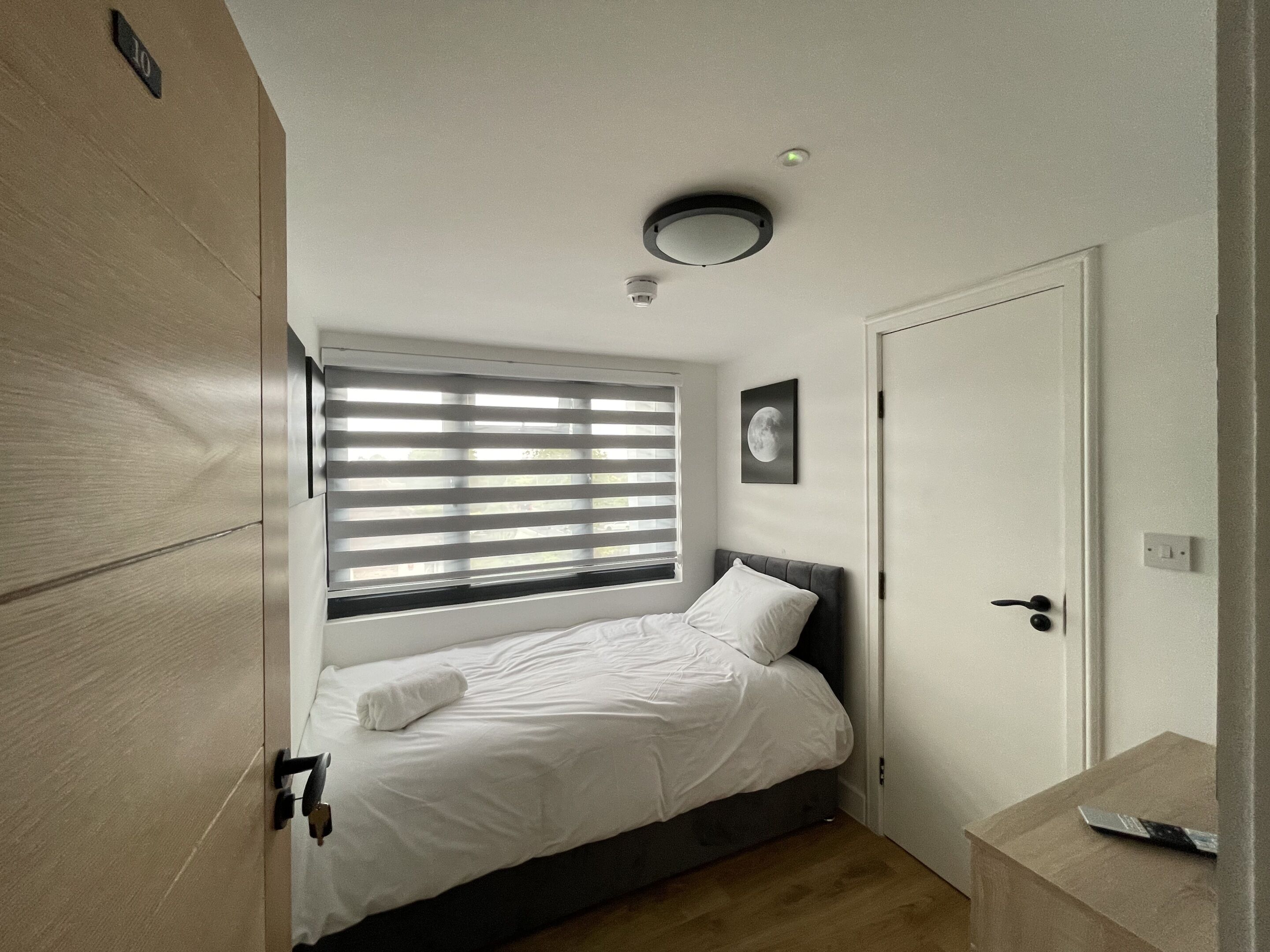 A bedroom with a bed, a window with blinds, a door, and a small bedside table.