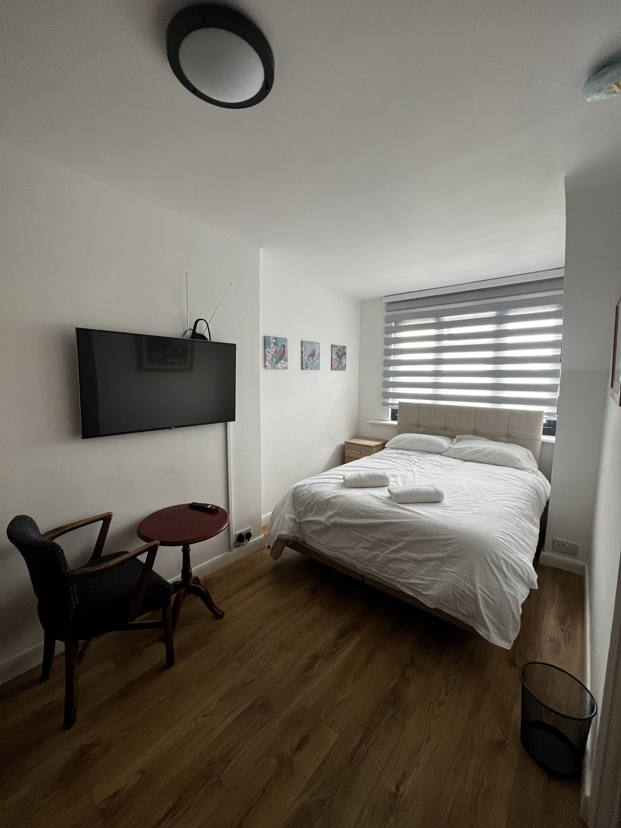 Un dormitorio con una cama, televisión montada en la pared, una mesita pequeña y una silla.