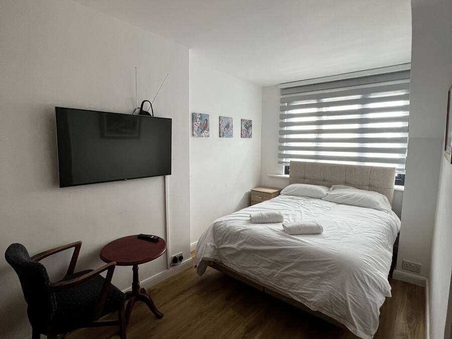Un dormitorio con una cama, televisión montada en la pared, una mesita pequeña y una silla.