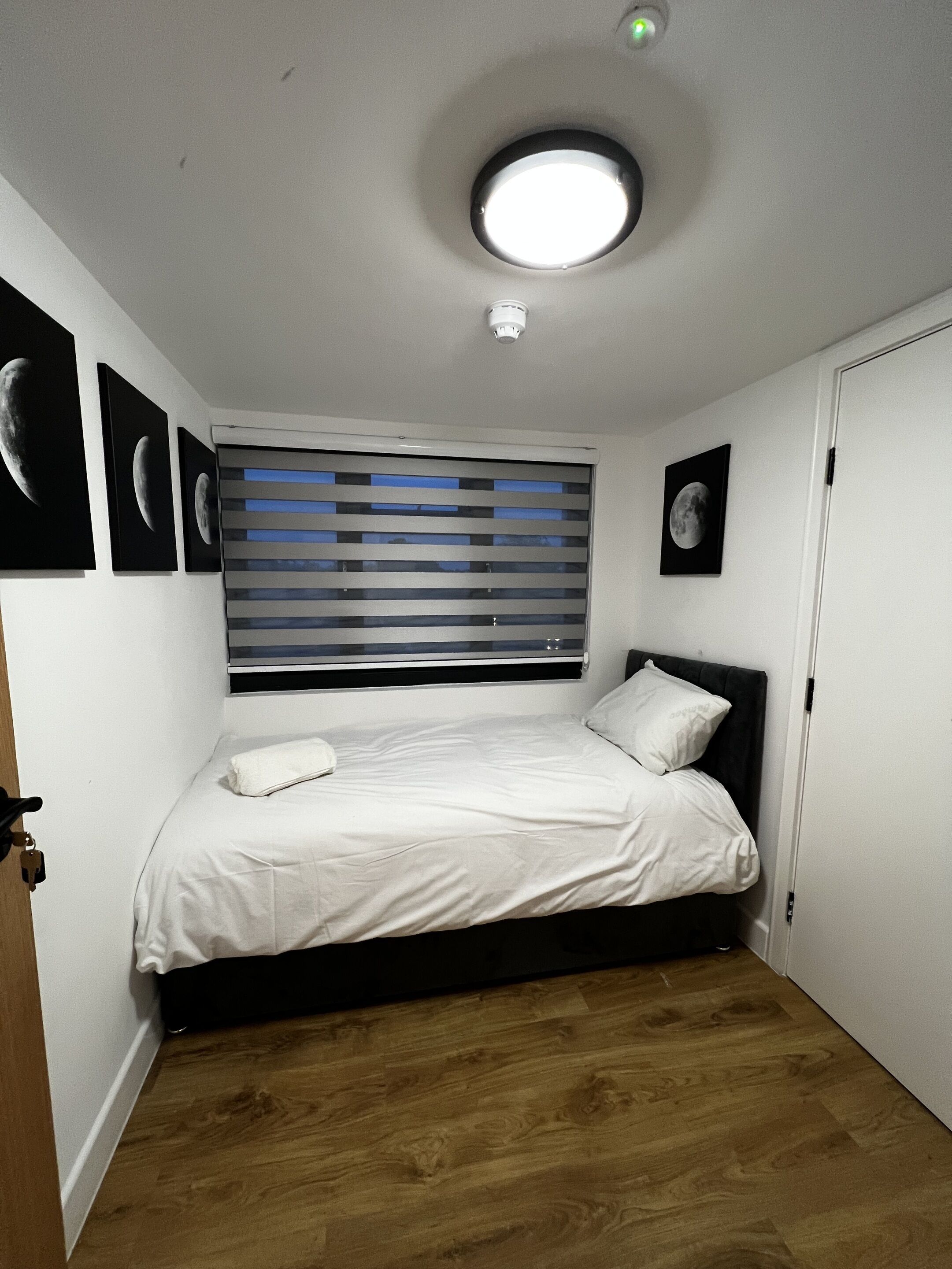 A bedroom with a bed, a window with blinds, a ceiling light, and wall-mounted pictures.