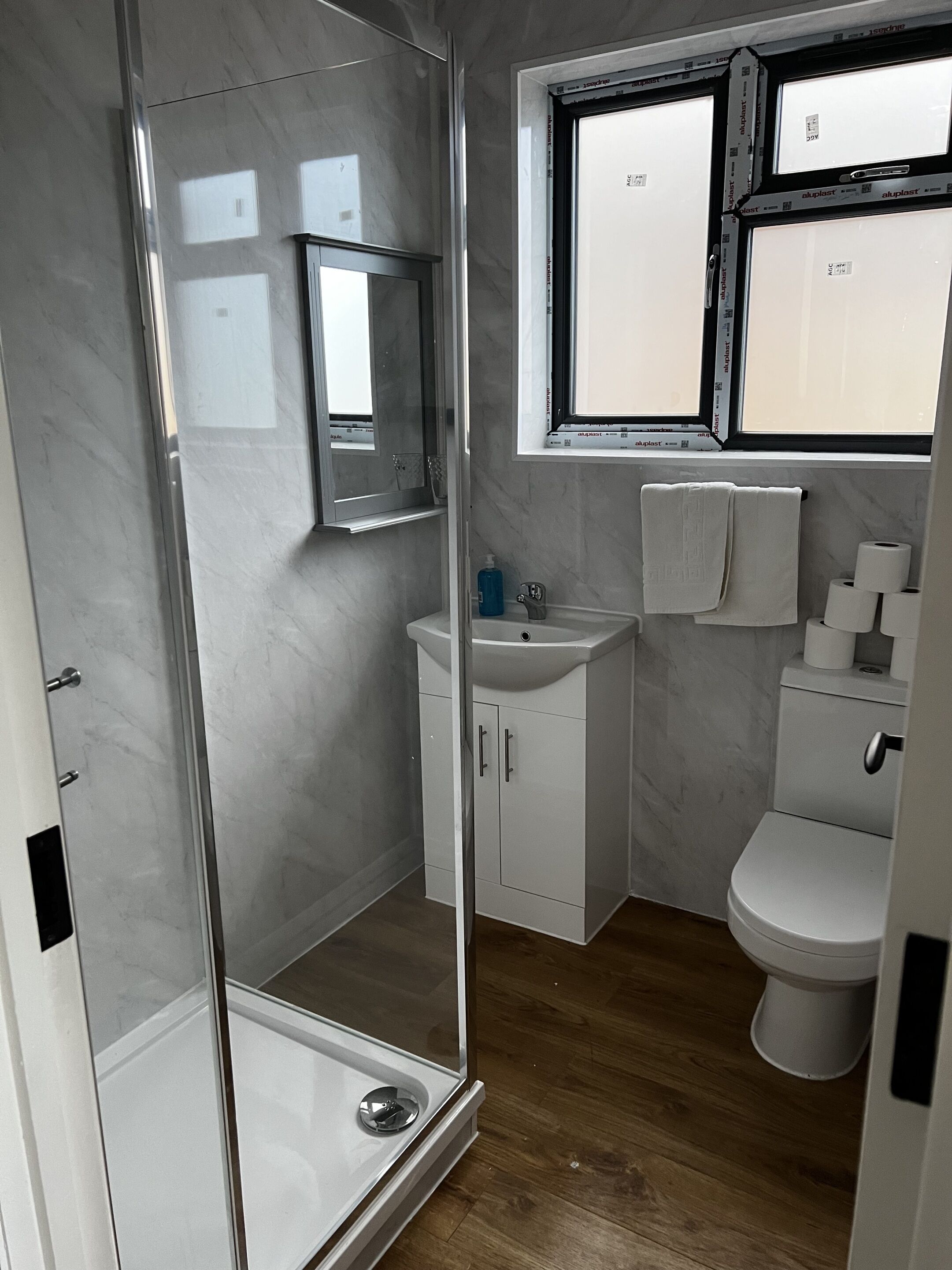 Un baño pequeño con cabina de ducha de vidrio, lavabo con espejo, inodoro y ventana sobre el lavabo.