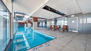 A modern indoor swimming pool with clear signage indicating shallow water depth, surrounded by glass walls and contemporary seating areas. - Modern & Stylish 2BR Apt - CBD - Free Parking (Adelaide)