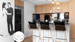 A modern kitchen with wooden cabinets, a black refrigerator, a microwave, and a coffee machine. There are three black bar stools with gold frames. - Modern & Stylish 2BR Apt - CBD - Free Parking (Adelaide)