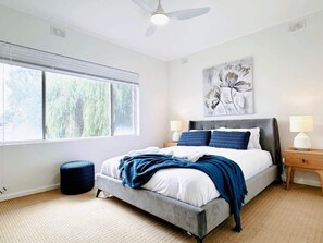 A modern bedroom with a large bed, two bedside tables, a ceiling fan, and a window with blinds. - In Between City, Sea & Airport - 2BR Vacation Home (Plympton)
