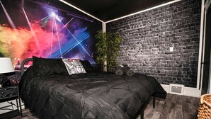 A bedroom with a black brick wall, a bed with a black duvet, a plant, and a wall-mounted speaker. - Cleveland’s Legends' Lair | Themed | Sleeps 4 (Cleveland)