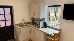 A compact kitchen with a sink, microwave, and cabinets, a small dining area with a chair, and a window with a view of a brick wall and a wooden fence. - Romantic King Bed Countryside Couples Cabin (Salisbury)