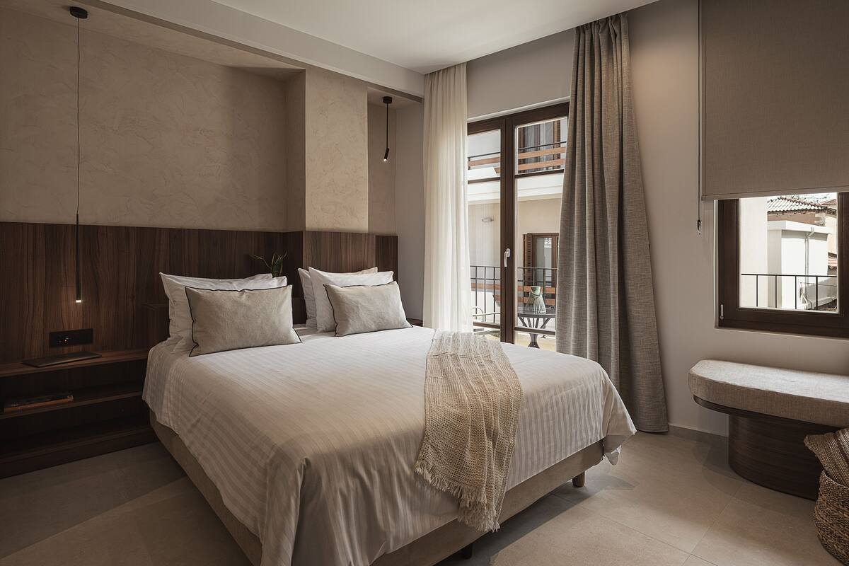 A bedroom with a large bed, a bedside table, a nightstand, a window with curtains, and a view of the outside balcony.