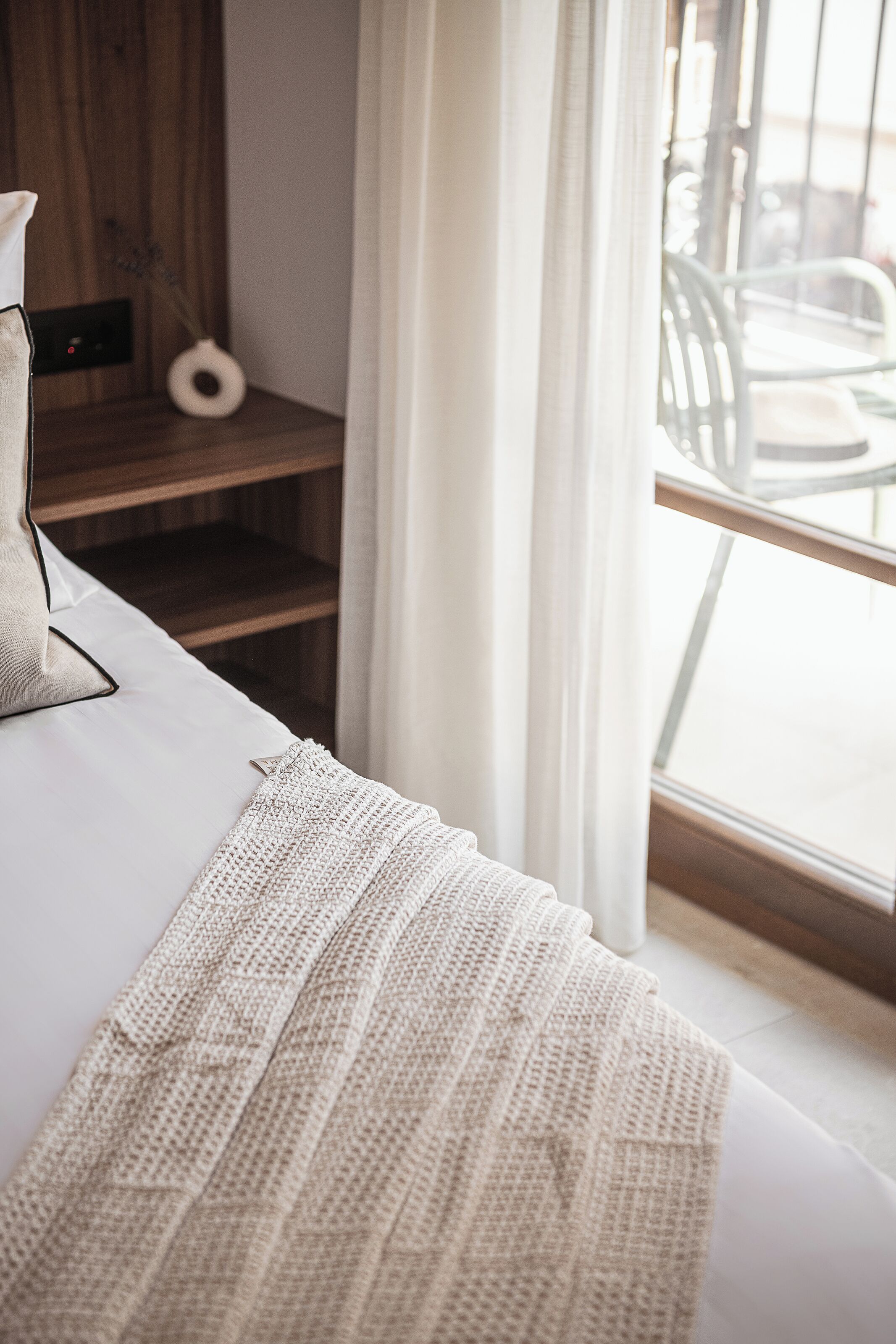 Ein ordentlich bezogenes Bett mit weißem Kopfkissen und einer Strickdecke, ein Holzregal mit einer Vase und ein Blick ins Freie durch ein mit Vorhängen drapiertes Fenster.