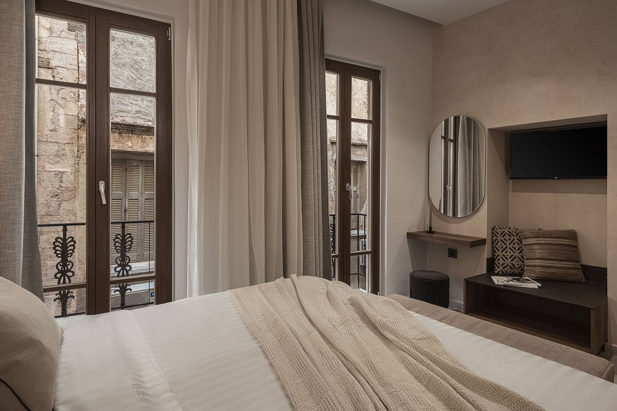 A bedroom with a large bed, a TV mounted on the wall, a window with curtains, and a brick wall.