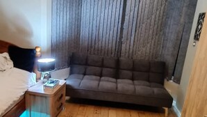 Une petite pièce chaleureuse, avec un plancher en bois, un canapé brun foncé, une table de chevet avec une lampe et une fenêtre avec des rideaux.